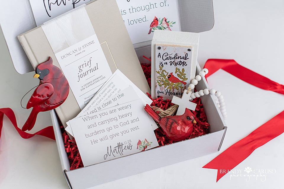 A sympathy gift box for grieving hearts includes a Cardinal glass ornament, Cardinal figurine, cross, table top Cardinal quote, guided grief journal
